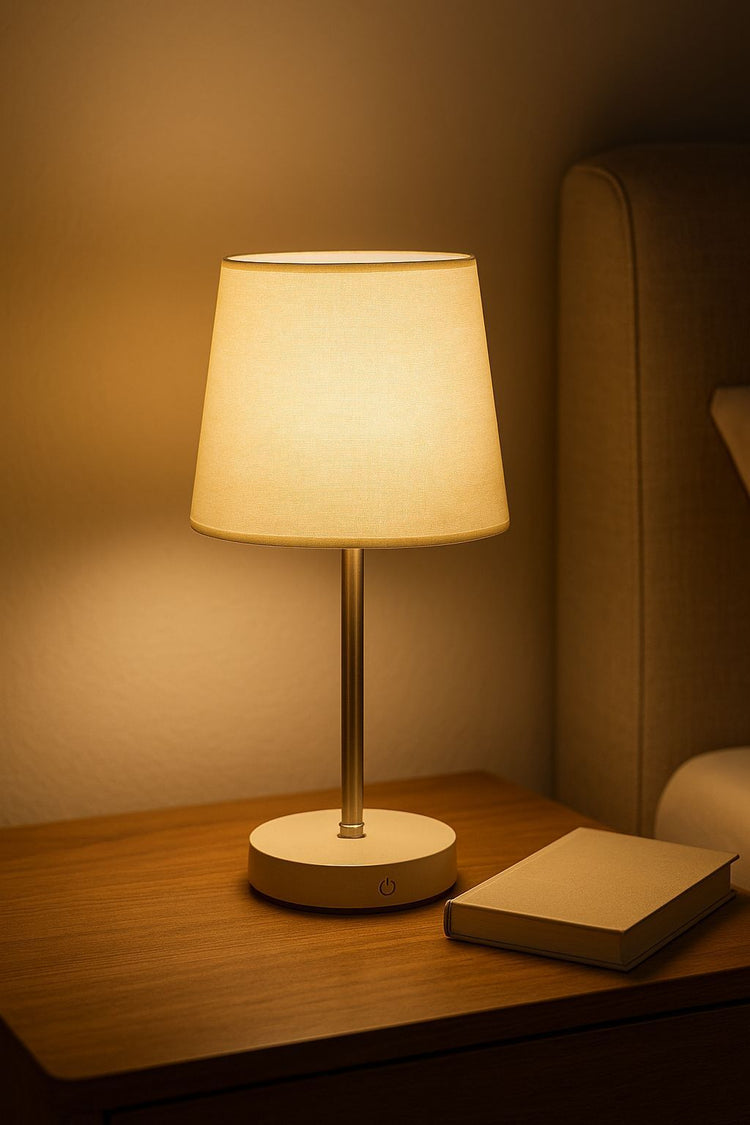 Lámpara de mesa con diseño clásico y pantalla de tela en tono neutro, colocada sobre una mesa de noche junto a una cama. Emite una luz cálida y suave, perfecta para lectura nocturna o crear un ambiente acogedor en el dormitorio.