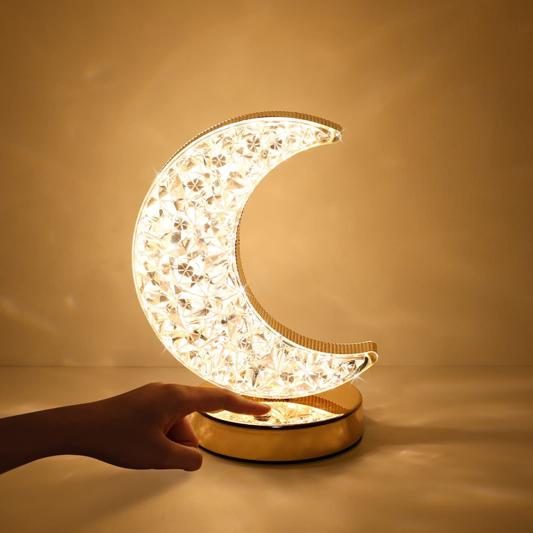 Lámpara decorativa con forma de luna creciente, fabricada con efecto de cristal brillante y montada sobre una base dorada. Ofrece luz cálida ideal para crear un ambiente acogedor en dormitorios, salas o espacios íntimos. Se activa fácilmente con un toque.