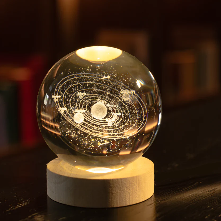 Lámpara decorativa con forma de globo de vidrio, con un diseño detallado del sistema solar grabado en su interior. La base de madera resalta la pieza mientras la luz cálida ilumina las estrellas y planetas, creando una atmósfera cósmica y única en cualquier espacio.