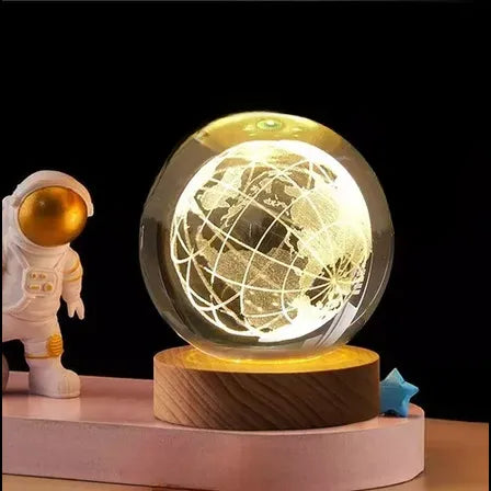 Lámpara decorativa LED con forma de globo terráqueo grabado en cristal 3D, montada sobre una base de madera que emite luz cálida. Acompañada de una figura de astronauta, es ideal para decorar escritorios, habitaciones o regalar a amantes de la geografía o el espacio.
