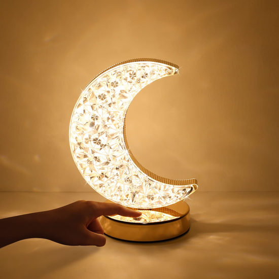 Lámpara decorativa con forma de luna creciente, fabricada con efecto de cristal brillante y montada sobre una base dorada. Ofrece luz cálida ideal para crear un ambiente acogedor en dormitorios, salas o espacios íntimos. Se activa fácilmente con un toque.