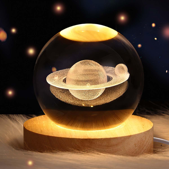 Lámpara decorativa con esfera de cristal que muestra un diseño en 3D del planeta Saturno con sus anillos, iluminada desde una base de madera que emite luz cálida. Ideal para decoración temática espacial, luz ambiental o como regalo original para fanáticos del universo.