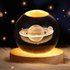 Lámpara decorativa con esfera de cristal que muestra un diseño en 3D del planeta Saturno con sus anillos, iluminada desde una base de madera que emite luz cálida. Ideal para decoración temática espacial, luz ambiental o como regalo original para fanáticos del universo.