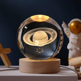 Esfera de cristal con diseño 3D de Saturno, iluminada desde una base de madera con luz cálida.
